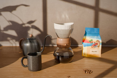 A glass pour-over brewer with a paper filter on a sunlit kitchen counter, next to a black gooseneck kettle, a steaming speckled mug of coffee and a bag of Santa Barbara Roasting Co. Peru N.G., with window and leaf shadows on the wall.