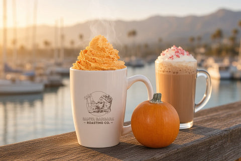 A pumpkin spice latte topped with orange whipped cream in a cream Santa Barbara Roasting Co. mug on a harbor railing at sunrise, beside a small pumpkin and a glass mug of peppermint mocha with whipped cream and crushed peppermint.