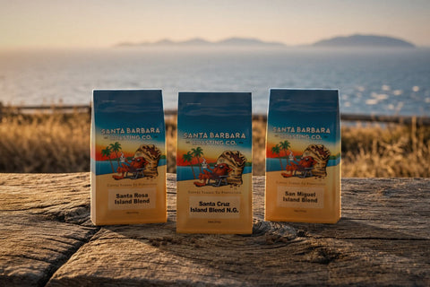 Three 12oz Santa Barbara Roasting Co. Naturally Grown Island Blend coffee bags, Santa Rosa, Santa Cruz and San Miguel, on a weathered wood table at golden hour with the Pacific and the Channel Islands softly blurred behind.