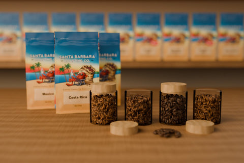 Glass jars of whole coffee beans with wooden lids on a cafe counter, next to a Santa Barbara Roasting Co. adventure pack, with shelves of coffee bags blurred behind.